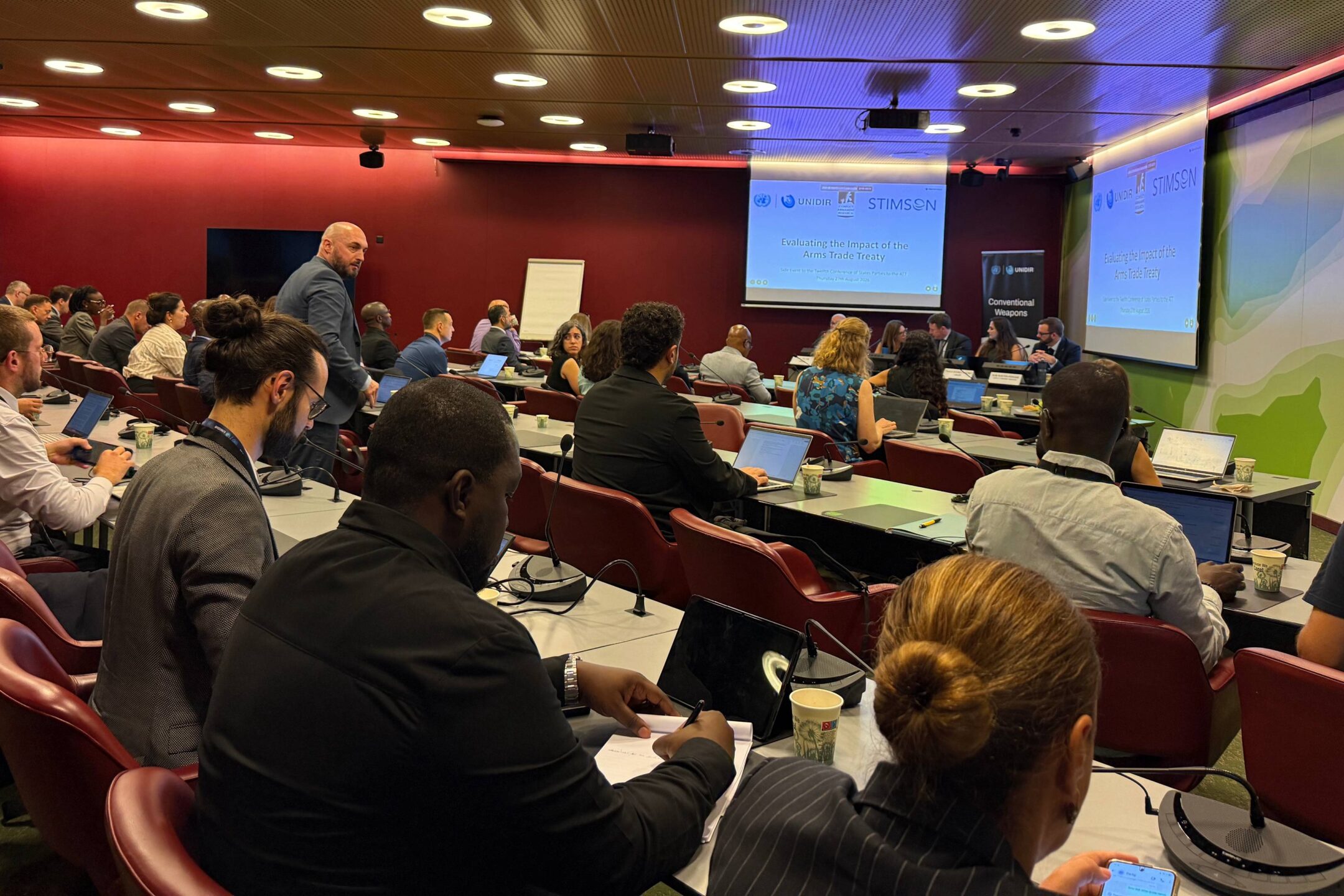 Participants seated at desks with laptops and notebooks at the side event “Evaluating the Impact of the Arms Trade Treaty”, held on the margins of the Twelfth Conference of States Parties to the Arms Trade Treaty in Geneva. The panel is visible at the front of the room.