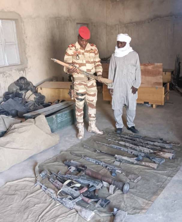 A Nigerien officer in desert camouflage and a red beret inspects a rifle beside a man in traditional dress, with collected rifles and weapon parts laid out on a tarpaulin in a storeroom, illustrating small arms collection efforts in Niger.