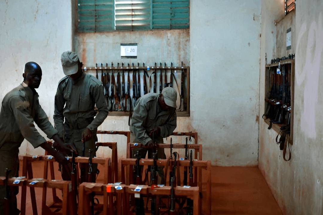 Three uniformed personnel handle rifles stored in numbered wooden racks inside a weapons storage room, with more rifles hung on wall racks, illustrating physical security and stockpile management.