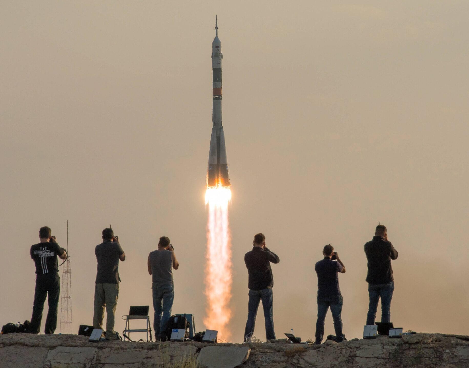 Six photographers, seen from behind on a concrete ledge, capture the Soyuz MS-01 rocket lifting off from the Baikonur Cosmodrome in Kazakhstan against a hazy sky. The scene illustrates the visibility of space activities that information-sharing under Article XI of the Outer Space Treaty seeks to extend.